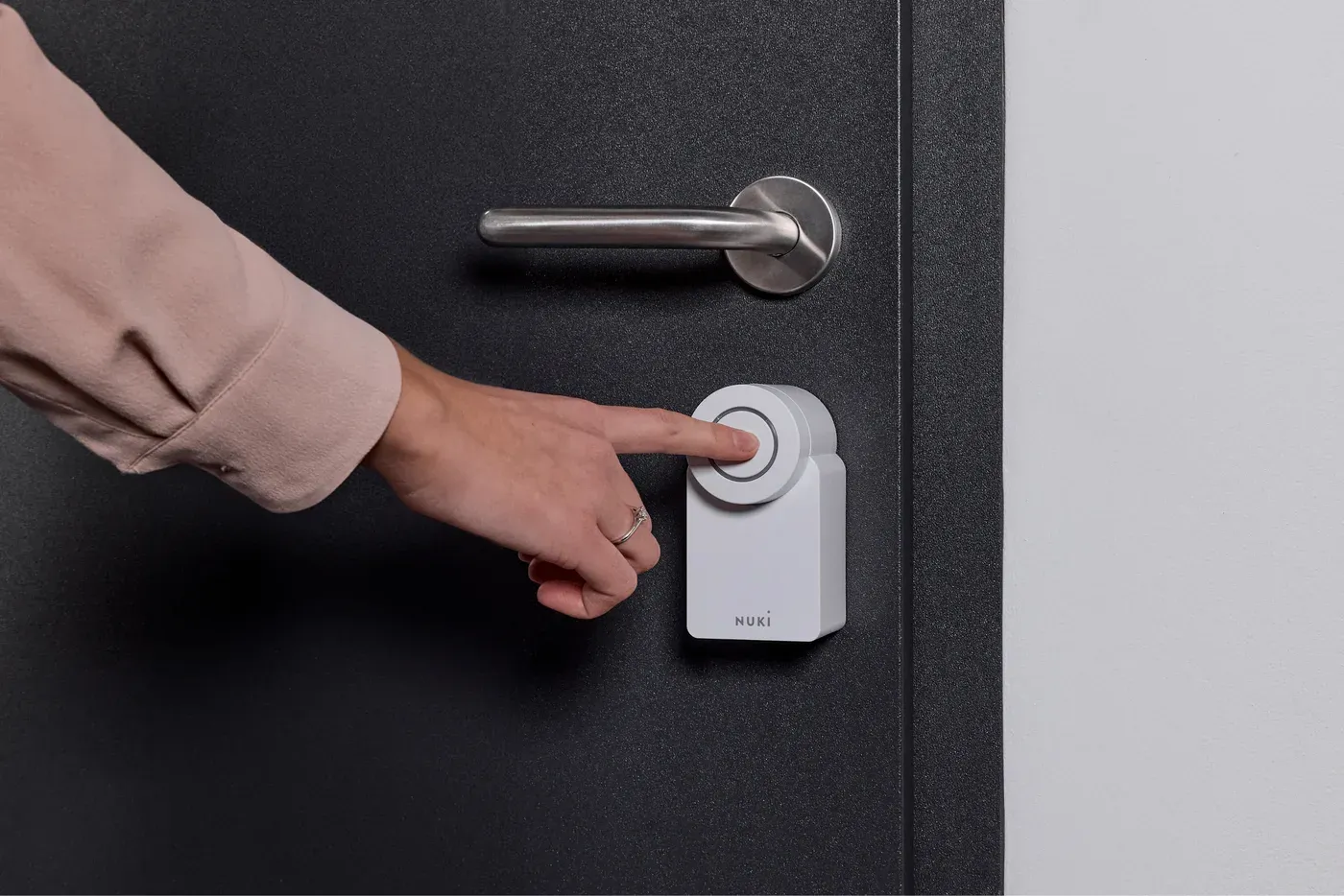 Une main appuie sur le bouton de la Nuki Smart Lock Go posée sous la poignée d'une porte sombre