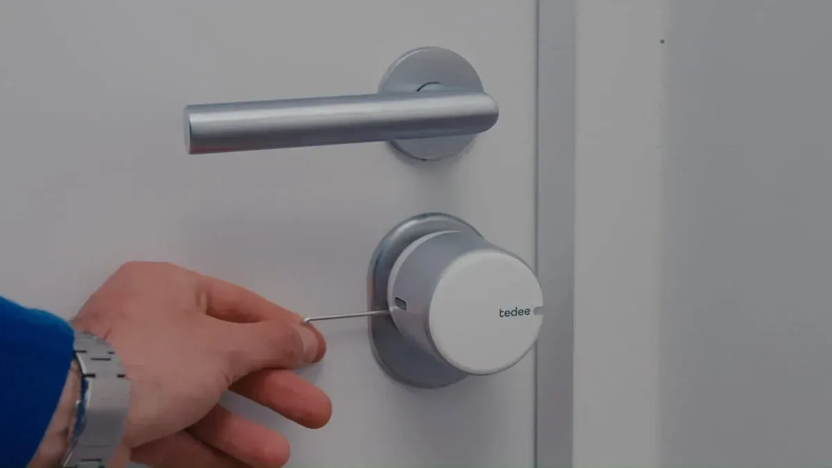 Une main serre à la clé Allen le Tedee Go argent posé sur le cylindre d'une porte blanche